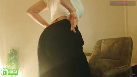 A video showing a woman with a light complexion wearing a white top and black pants, standing with her hands on her hips, in a dimly lit room with a brown chair and green decor in the background.