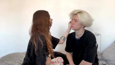 Video of a young woman with long, straight brown hair in a black shirt, touching a young man with platinum blonde hair wearing a black t-shirt. They sit on a bed with white walls in the background.