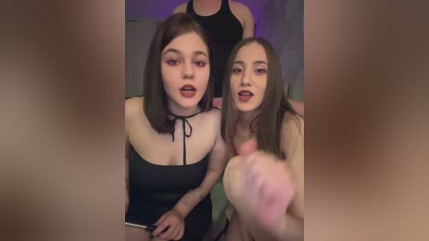 Video of two young women with fair skin and shoulder-length brown hair, wearing black tank tops, sitting on a couch; blurred background.