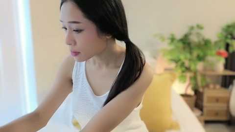 Video of a young East Asian woman with long black hair in a white tank top, sitting thoughtfully on a bed, in a softly lit room with green plants and wooden furniture.
