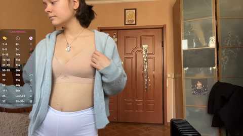 Video of a young Asian woman in a beige bra, white pants, and light blue cardigan, standing in a cozy, beige-walled room with a wooden door and glass cabinet.