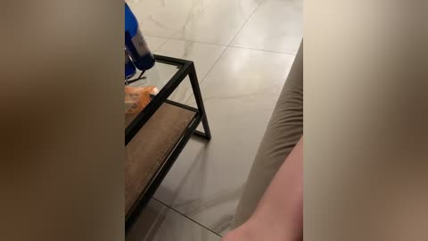 A video of a beige-tiled bathroom with a black metal tray holding a blue bottle, orange bread, and a white bowl. A person\u2019s bare leg is visible in the bottom right corner.