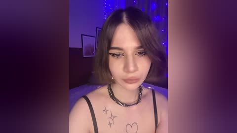 Video of a young woman with straight, shoulder-length brown hair, wearing a black spaghetti-strap top, adorned with star tattoos on her collarbone. She has a neutral expression under dramatic winged eyeliner. The background features dim blue LED lighting and framed photos.