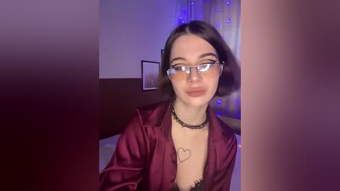 Video of a young woman with fair skin, dark hair, and glasses, wearing a burgundy satin robe, black choker, and revealing black lingerie, sitting in a dimly lit room with purple fairy lights.