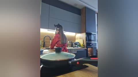 A video of a woman in a red robe cooking on a stove in a modern kitchen with black and wood accents, wearing a black cat mask.