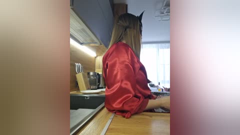 Video of a woman in a red satin robe, sitting on a wooden kitchen counter, facing away, with a knife block and kitchen utensils in the background.