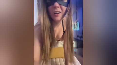 Video of a blonde woman wearing a black mask, low-cut white dress, and yellow belt, indoors with blurred background.