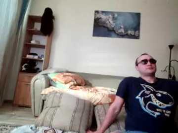 A video of a man in a dark T-shirt, sunglasses, and jeans, sitting on a beige couch with a striped blanket, in a cluttered, beige-walled room with a wooden bookshelf, a framed ocean landscape, and a hanging jacket.