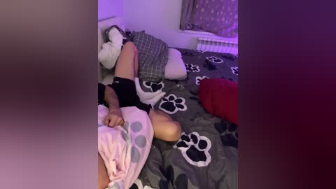 Video of a woman in a messy bedroom, lying on a bed with a paw-printed duvet, wearing a black top and pink pajama shorts, with a stuffed bear beside her.