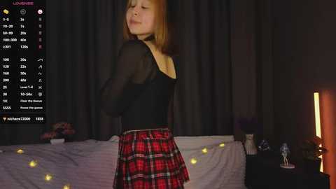 Video of a young woman with fair skin and red hair, wearing a black top and red plaid skirt, standing in a dimly lit bedroom with dark curtains, digital clock, and a bed.