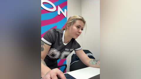 Video of a tattooed woman in a grey sports jersey, leaning over a white table in a colorful, striped background with blue and pink tones.