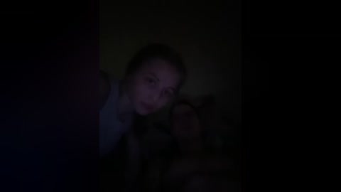 A dimly lit video of a woman with dark hair, wearing a blue top, and a young girl with blonde hair, both with serious expressions, in a dark, shadowy room.