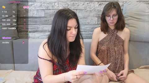 Video of two women: one in a red dress, the other in a leopard-print dress, reading a magazine on a gray sofa against a rustic wooden wall.