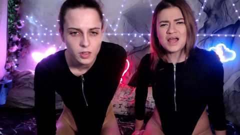 Video of two young women with fair skin and brown hair, wearing black zip-up tops, sitting close on a bed with a gray and white patterned blanket. The background features a mural, fairy lights, and a neon cloud sign, creating a cozy, intimate atmosphere.