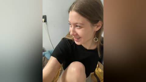 Video of a young woman with light skin, brown hair, and a black t-shirt, smiling while seated indoors. She wears gold hoop earrings. Background features a gray wall with a black power outlet and a yellow blanket.