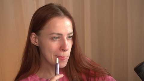 Video of a young woman with straight, long, reddish-brown hair, fair skin, and green eyes, applying makeup with a dark eyeliner. She wears a pink shirt, and the background is a beige curtain.