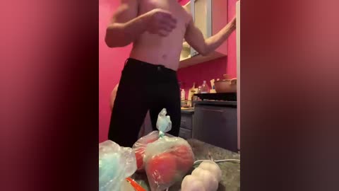 Video of a topless man with a muscular build, wearing black pants, standing in a kitchen with pink walls, holding a blue plastic bag containing fruits and vegetables.