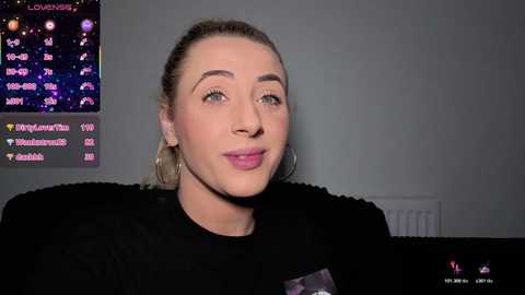 Video of a light-skinned woman with blonde hair in a ponytail, wearing a black top, large hoop earrings, and light makeup, seated indoors with a wall-mounted TV displaying a star map and social media icons.