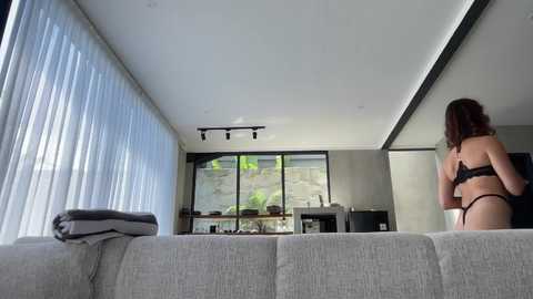 Video of a modern living room with large windows, grey couch, woman in black lingerie, flat-screen TV, minimalist decor, and natural light.