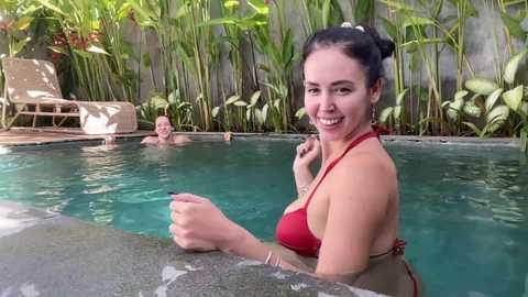Video of a smiling woman with dark hair in a red bikini, standing in a clear pool with lush green plants and a man lounging nearby.