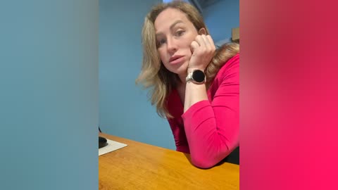 Video of a light-skinned woman with wavy blonde hair, wearing a pink top, resting her chin on her hand at a wooden desk, surrounded by a blue wall and a pink frame.