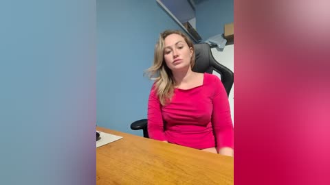 Video of a blonde woman with fair skin, wearing a bright pink long-sleeve top, sitting at a wooden desk, eyes closed, in a dimly lit office with blue walls.