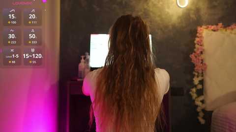 Video of a woman with long, wavy, light brown hair, wearing a white top, standing in front of a computer screen displaying a weather forecast and temperature. Background includes a floral arrangement and dim lighting.