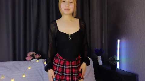 Video of a smiling, fair-skinned woman with blonde hair in a plaid skirt, black top, and sheer sleeves, standing in a dimly lit room with gray curtains, white bed, and fairy lights.