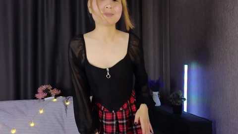 Video of a fair-skinned woman with short blonde hair, wearing a black sheer top and red plaid skirt, standing in a dimly lit room with a bed and purple flowers.