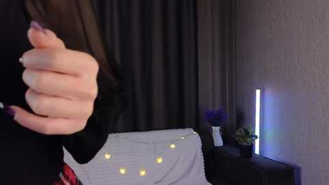 Video of a woman's hand in black clothing, holding a small object, against a background of a bed with white and yellow string lights, dark curtains, and a purple potted plant.