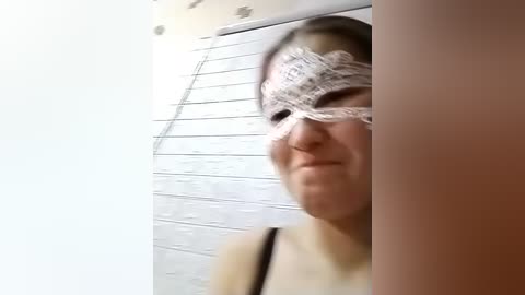 Video of a person wearing a white lace masquerade mask, with a faint smile. The background features a white, tiled wall with a vertical window. The image has a soft focus.