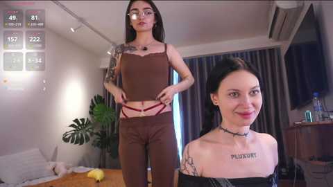 Video of a young woman with long black hair, glasses, and tattoos, wearing brown lingerie, standing in a modern, minimalist bedroom.