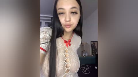 Video of a young woman with long black hair, fair skin, and large breasts, wearing a sheer white lace top with red bows. Background shows a cluttered room with a window and framed pictures.