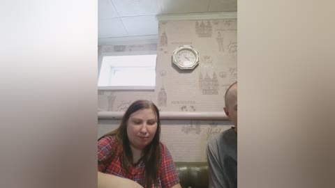 Video of a middle-aged woman with long brown hair, wearing a red plaid shirt, sitting on a green leather couch in a room with beige wallpaper featuring historic building illustrations and a round clock.