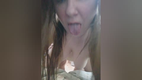 A video of a young, fair-skinned woman with long, wet hair, sticking out her tongue, partially nude, with her breasts visible, on a bed with a beige blanket.