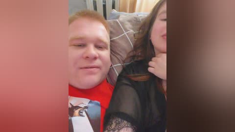 A video of a close-up selfie featuring a man with red hair in a red T-shirt, and a woman with long brown hair in a black lace top, both resting on a bed with patterned pillows.