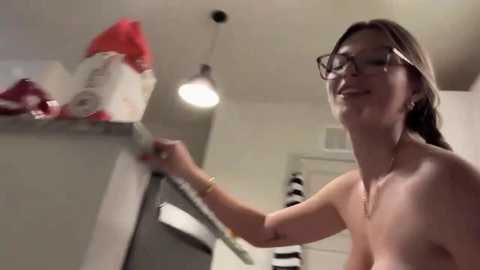 Video of a topless woman with glasses and light brown hair, cleaning a countertop with a sponge, in a modern kitchen with white cabinets and pendant lights.