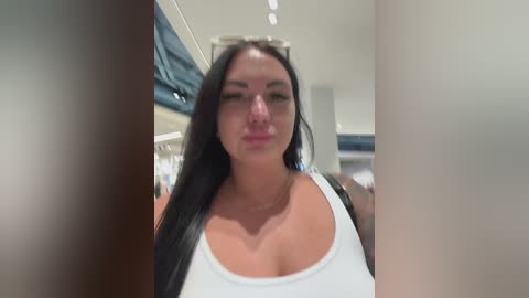 A video of a fair-skinned woman with long black hair, wearing a white tank top, standing in a modern, brightly lit indoor setting, possibly a mall or store.