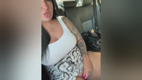 Video of a tattooed woman in a white tank top, snake-patterned skirt, sitting in a car, with a blurred background.