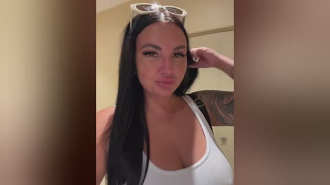 Video of a fair-skinned, tattooed woman with long black hair, wearing a white tank top, sunglasses on her head, and a confident expression.