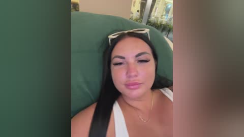 Video of a young woman with long black hair, wearing sunglasses, a white top, and a gold necklace, lying on a green couch.