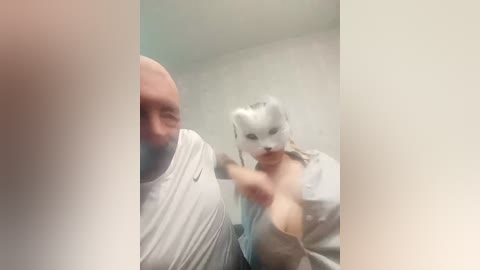 A blurred video shows a bald man in a white shirt and a white-masked woman holding a pink object, possibly a phone, in a dimly lit room with a beige wall.