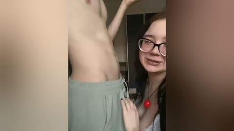 Video of a smiling, glasses-wearing woman with long brown hair and a red necklace, hugging a shirtless man with fair skin and toned abs. They're in a dimly-lit room, with a mirror and blurred background.