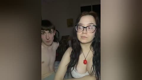 A video of a young, pale-skinned woman with long black hair, wearing glasses and a red necklace, sitting on a bed in a dimly lit room. A shirtless young man with messy brown hair stands behind her, looking curious.