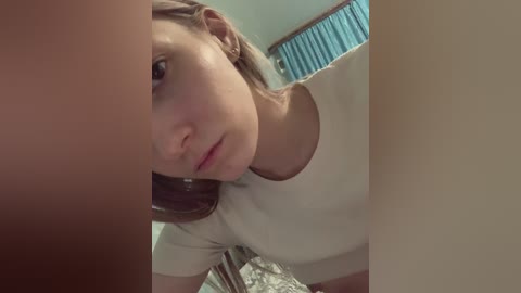 A video of a young woman with fair skin and brown hair, wearing a white top, taken from a low angle, showing her face and shoulders. The background includes a teal curtain and a bed with a patterned blanket.