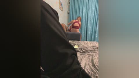 Video of a woman with auburn hair in a red bra and black pants sitting on a bed with a gray blanket, adjusting her hair in front of a mirror with turquoise curtains.