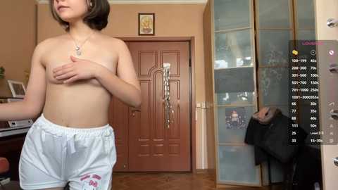 Video of a young, topless woman with short brown hair, wearing white shorts, standing in a modern, beige-tiled living room.