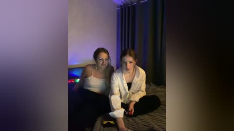 Video of two women playing a video game in a dimly-lit room. One woman wears a white blouse and black pants, the other a white robe.