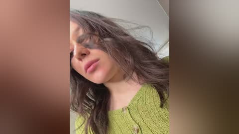 Video of a young woman with wavy brown hair, wearing a green sweater, slightly blurred, with a blurry background, and a blurred man's face in the foreground.