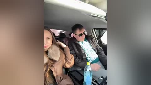 Video of a young woman with long brown hair, brown coat, and fur-trimmed hood, sitting next to a young man with short dark hair, sunglasses, and a green t-shirt, inside a car.